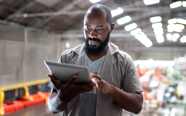 A man standing in a warehouse uses a tablet, focusing on the screen with equipment and shelves visible in the blurred background.