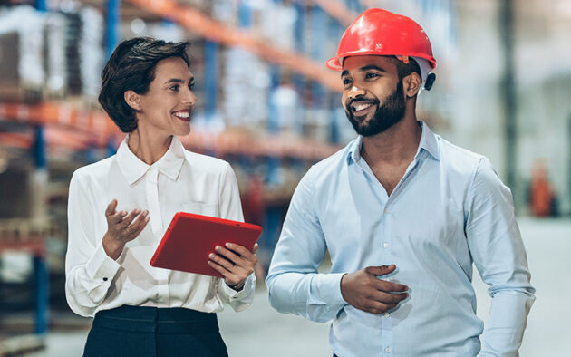 A woman holding a tablet talks to a man wearing a red hard hat as they walk through a warehouse or industrial facility.
