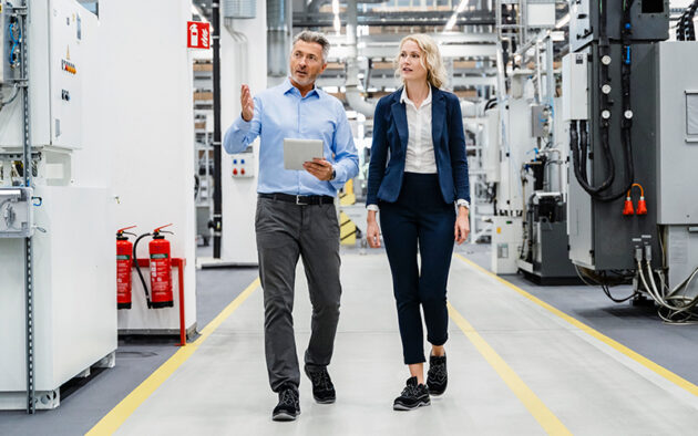 Two people in business attire walk and talk in a modern factory or industrial facility, surrounded by machinery and equipment.