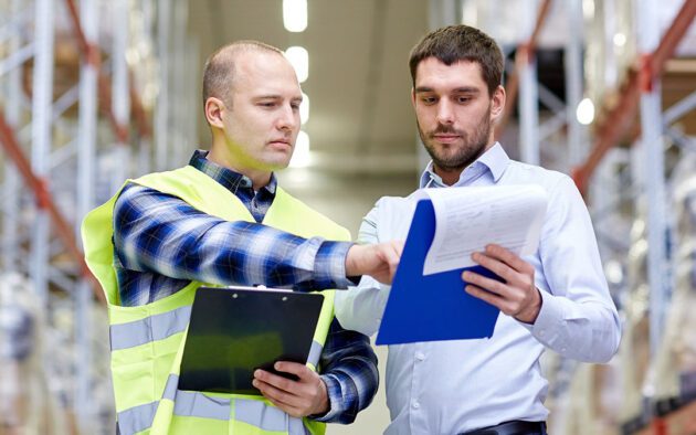 Two men, one in a reflective vest and another in a dress shirt, both looking at clipboards while inside a warehouse