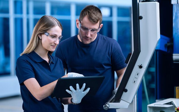 Two people wearing safety glasses and blue shirts review information on a digital tablet in an industrial or manufacturing setting.