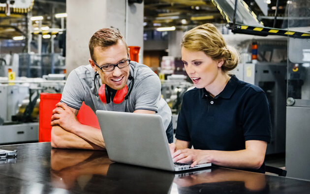 Two workers in a factory setting look at a laptop on a table; one is typing while the other watches and smiles. Machinery and equipment are visible in the background.