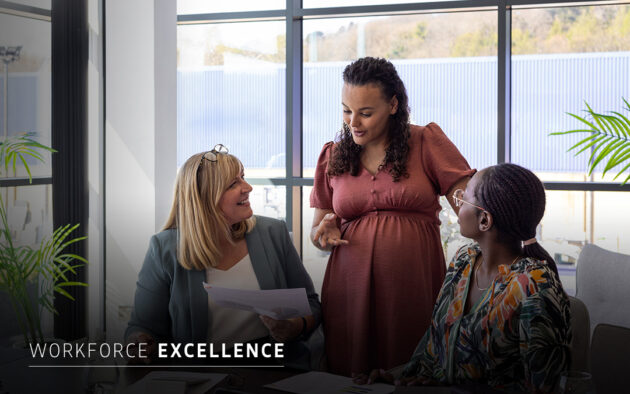 Three women in business attire are having a discussion at a meeting table in a modern office with large windows. Text reads “Workforce Excellence.”.