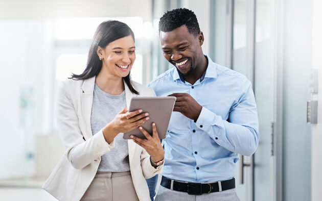 Two professionally dressed people standing in a bright office hallway, smiling and looking at a tablet together.