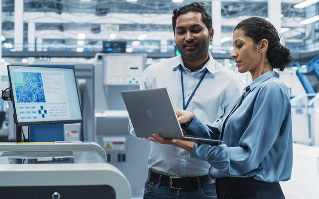 Two professionals in a factory setting review data on a laptop while standing near industrial equipment and a monitor displaying technical information.