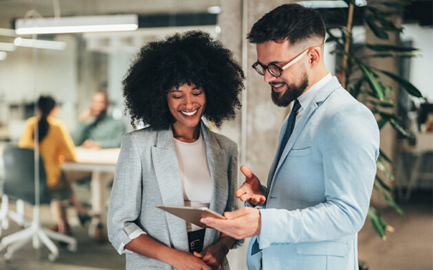 Two professionally dressed people stand in an office, smiling and looking at a tablet, with colleagues working in the background.