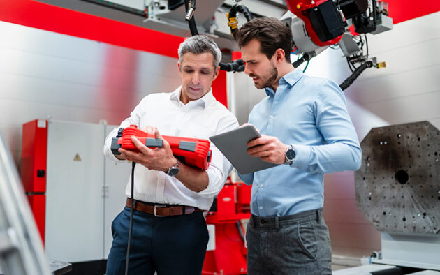 Two men in business attire examine a red industrial device and review information on a tablet in a modern manufacturing facility.