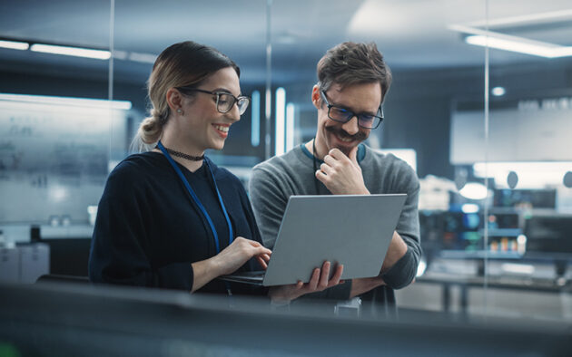 Two people wearing glasses stand together in an office, looking at a laptop and smiling, surrounded by modern technology equipment.